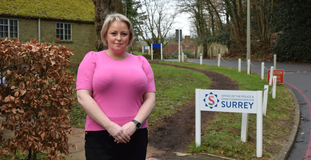 Commissioner Lisa Townsend standing in a pink top and smart trousers outside in front of a white sign that says Office of the Police and Crime Commissioner for Surrey