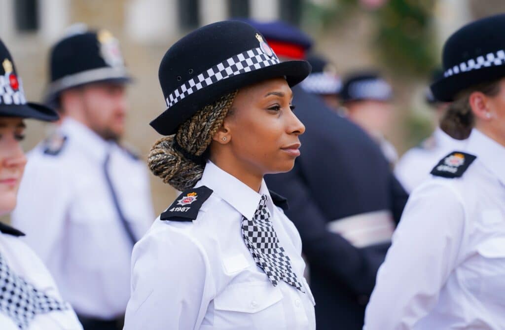 young black female police officer with subtle smile in smart black and white formal uniform and hat, as she stands with other new recruits to Surrey Police in 2022.