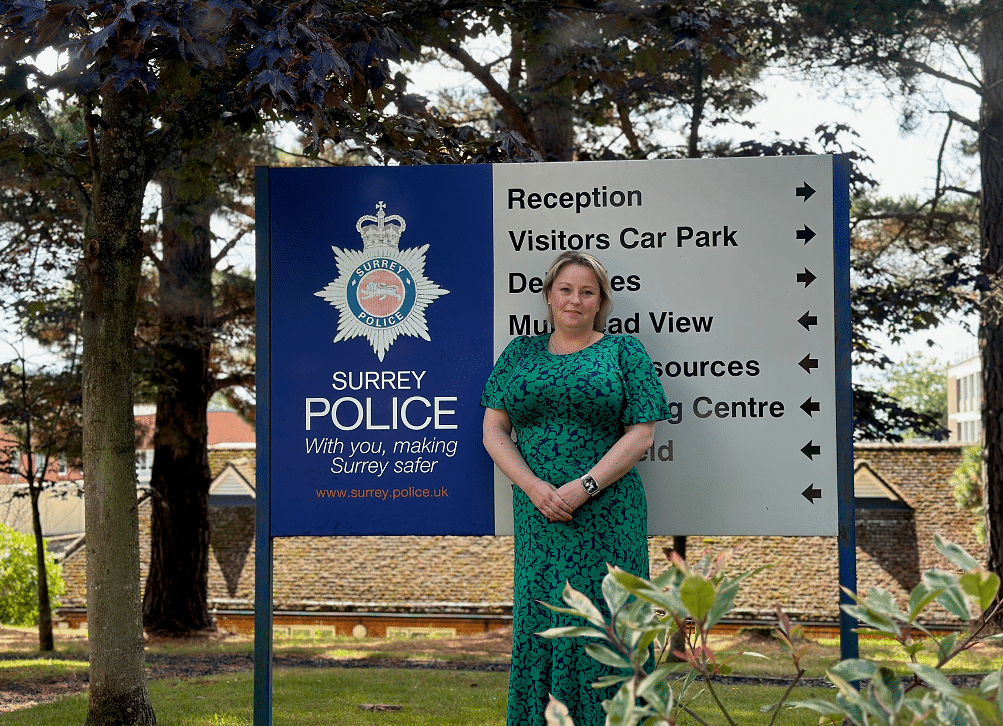 A Comissária de Polícia e Crime de Surrey, Lisa Townsend, em frente a uma placa de direção no QG da Polícia de Surrey, com árvores e edifícios ao fundo.