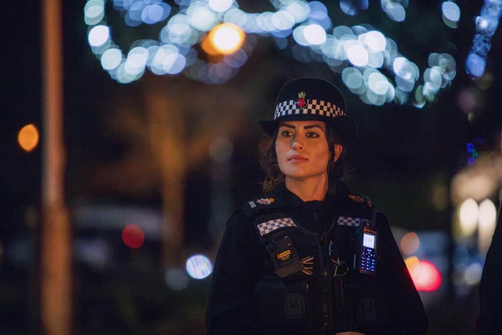 smart female police officer walking at night in full uniform. Glittering Christmas lights with a blue hue can be seen above the street behind her 