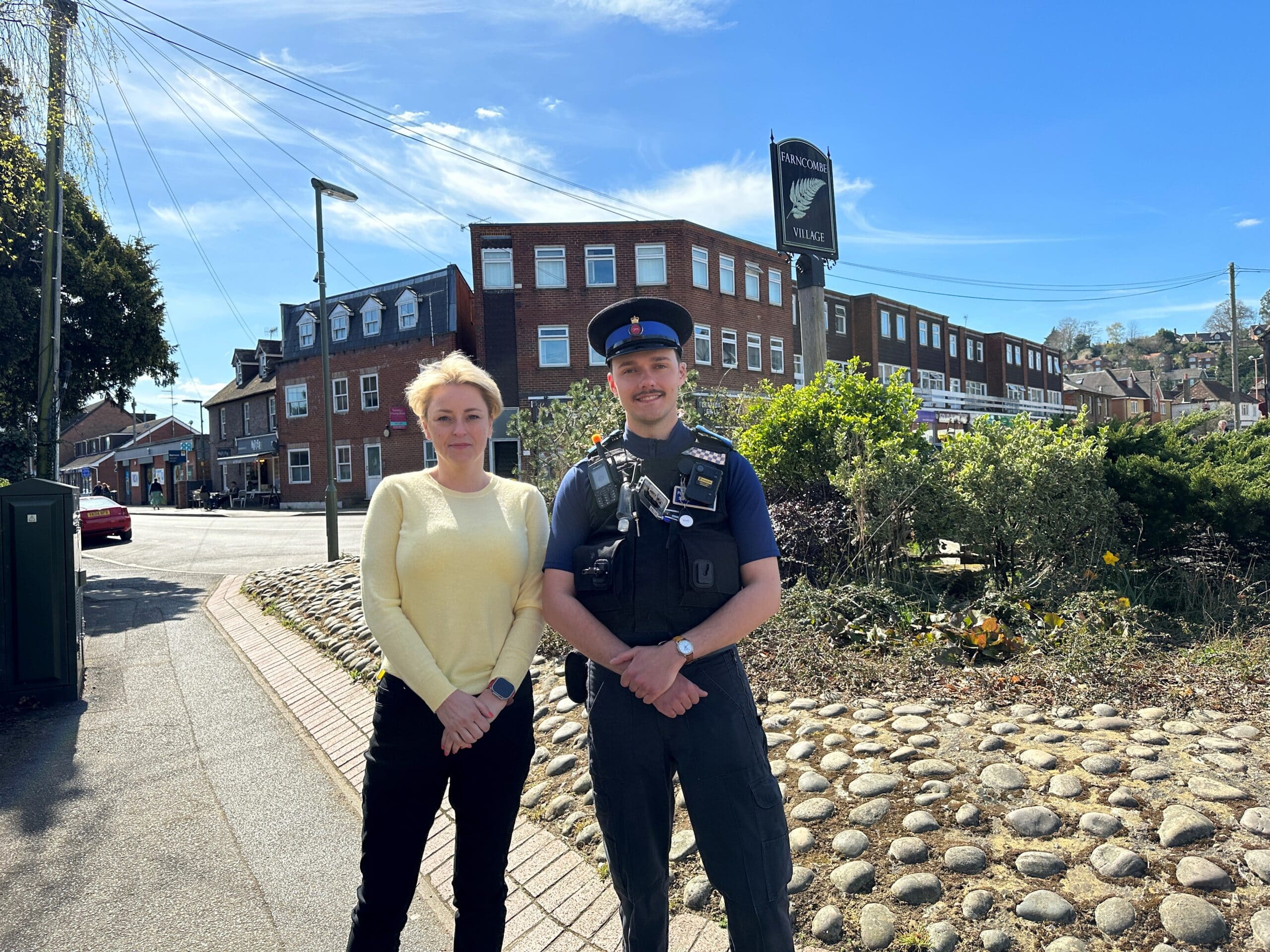 Two people stand together with their hands clasped looking at the camera. To the left of the image is Lisa Townsend, wearing a yellow jumper and black trousers. To the right is PCSO John Savage, who wears full uniform and a hat. They are standing in Farncombe centre, and a sign above their heads reads 'Farncombe'.