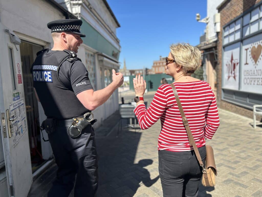 Two people are pictured walking along a street. Their backs are to the camera and they have partially turned and are talking to each other animatedly. On the left is a police officer in full uniform, and on the right is a blonde woman in a red and white long-sleeved top. To their right is a branch of Pret.
