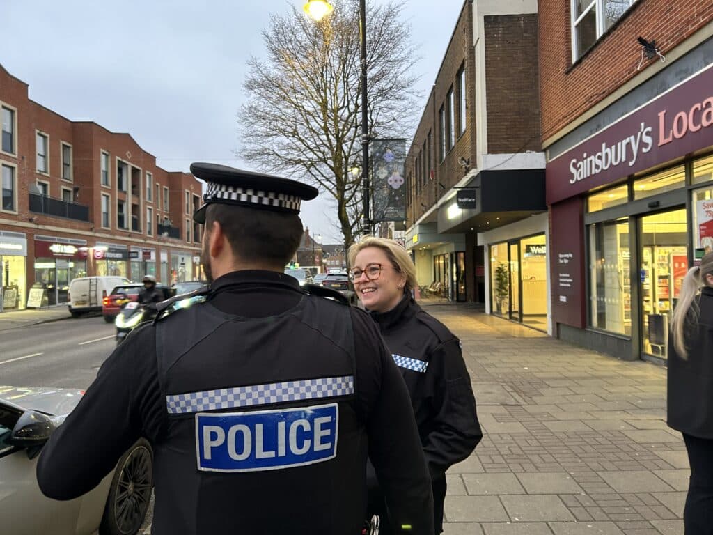 A police officer converses with the Police and Crime Commissioner for Surrey on a city street, both appearing engaged in discussion.