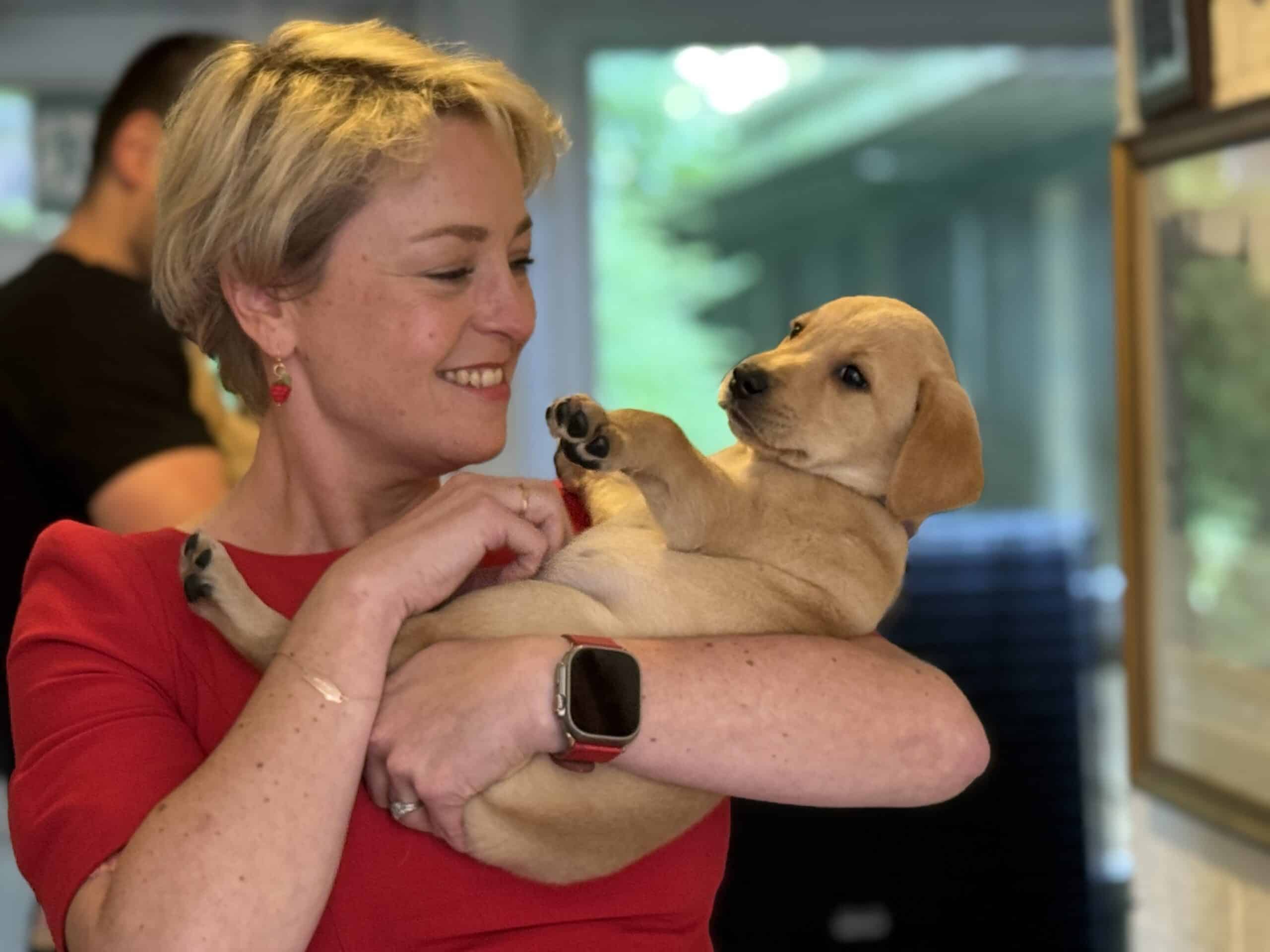 The picture shows Commissioner Lisa Townsend, who is wearing a red dress, red earrings, and has short blonde hair. She is cradling a golden Labrador puppy in her arms and smiling at it.