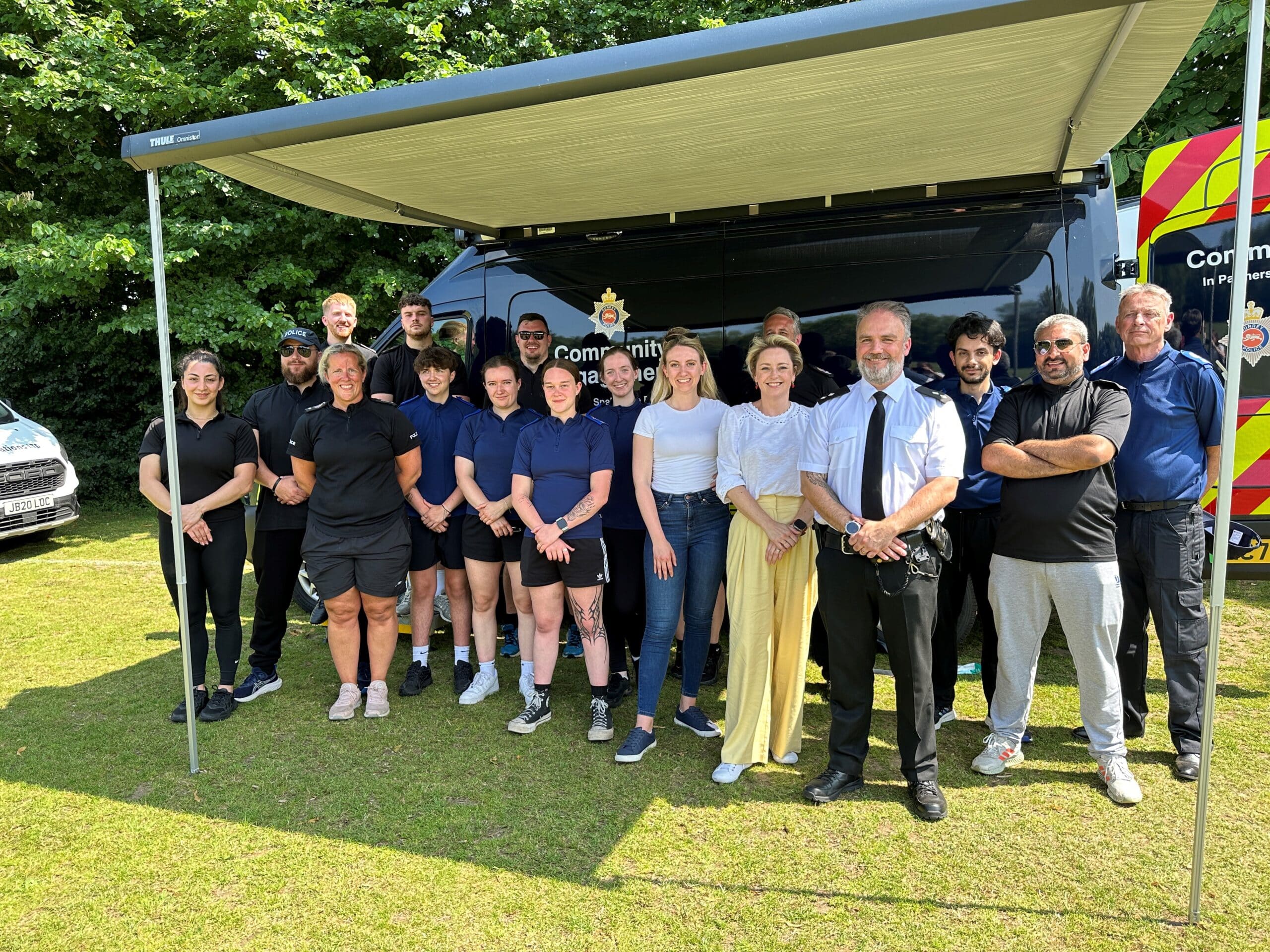 The picture shows a large group of people standing in front of a van with an awning over it. Among the group are people wearing Surrey Police-branded outfits. At the forefront stand Commissioner Lisa Townsend, who wears yellow trousers, and Deputy Commissioner Ellie Vesey-Thompson, who is in jeans. To their left is Borough Commander Matt Walton, who wears a shirt and tie. All of those in the image are standing on a field. They are all looking at the camera and smiling.
