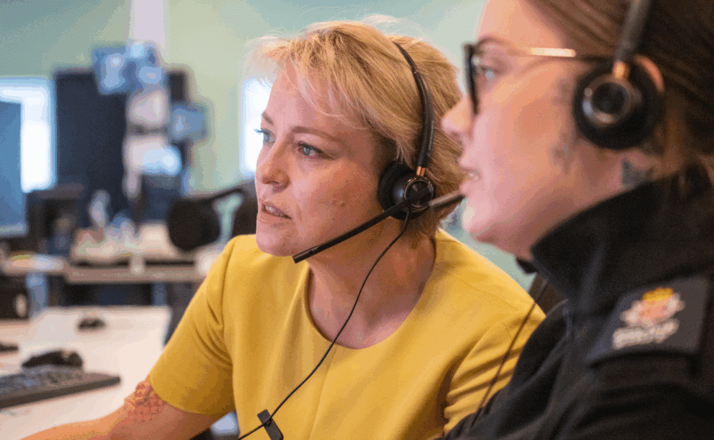 Two women sit beside each other. They are both wearing headsets and looking at a computer. The woman on the right, Commissioner Lisa Townsend, wears a yellow top. She is gazing intently at the computer screen. The woman on the right, who is in Surrey Police uniform and wears glasses, also looks at the screen. The image was taken at the Surrey Police contact centre in Guildford.