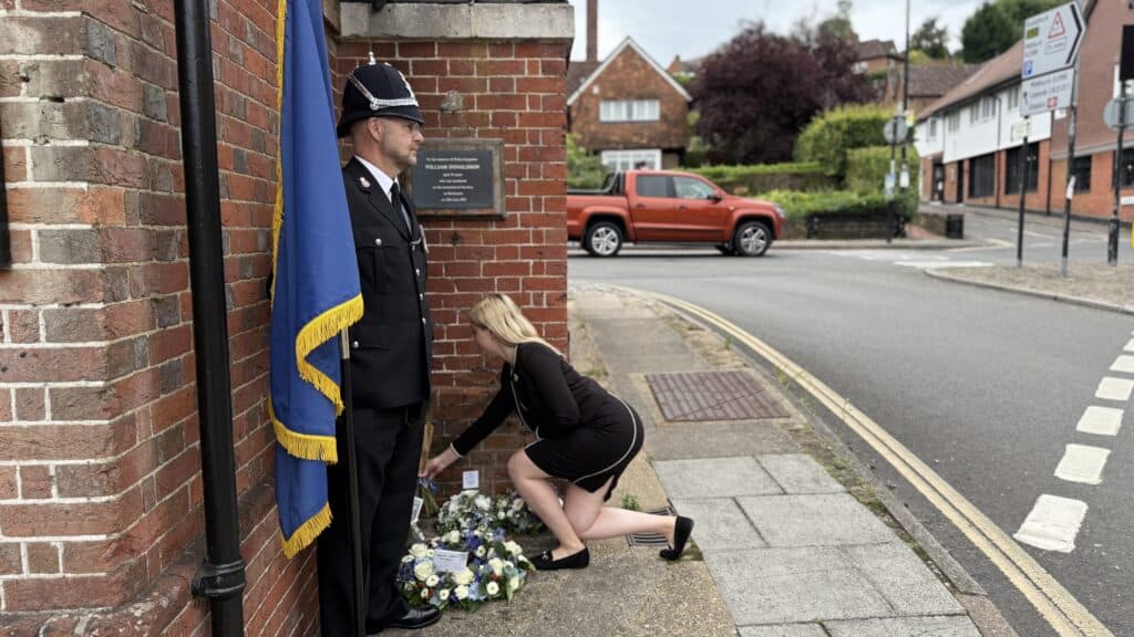 Image shows an officer in formal uniform holding a flag. Beside him, Deputy Commissioner Ellie Vesey-Thompson - a blonde woman wearing a black dress - kneels to place flowers beside a building.
