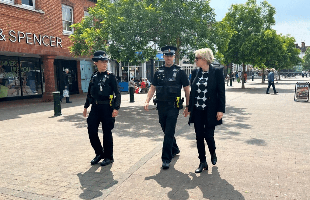 The image shows Police and Crime Commissioner Lisa Townsend walking through a town centre with two police officers in uniform. Lisa, who has blonde hair, stands on the right of the image. She wears a black jacket and trousers. The officers, one male and one female, are wearing their uniforms and police hats, and they are in the middle of a conversation with Lisa as they all walk past a branch of Marks and Spencer.
