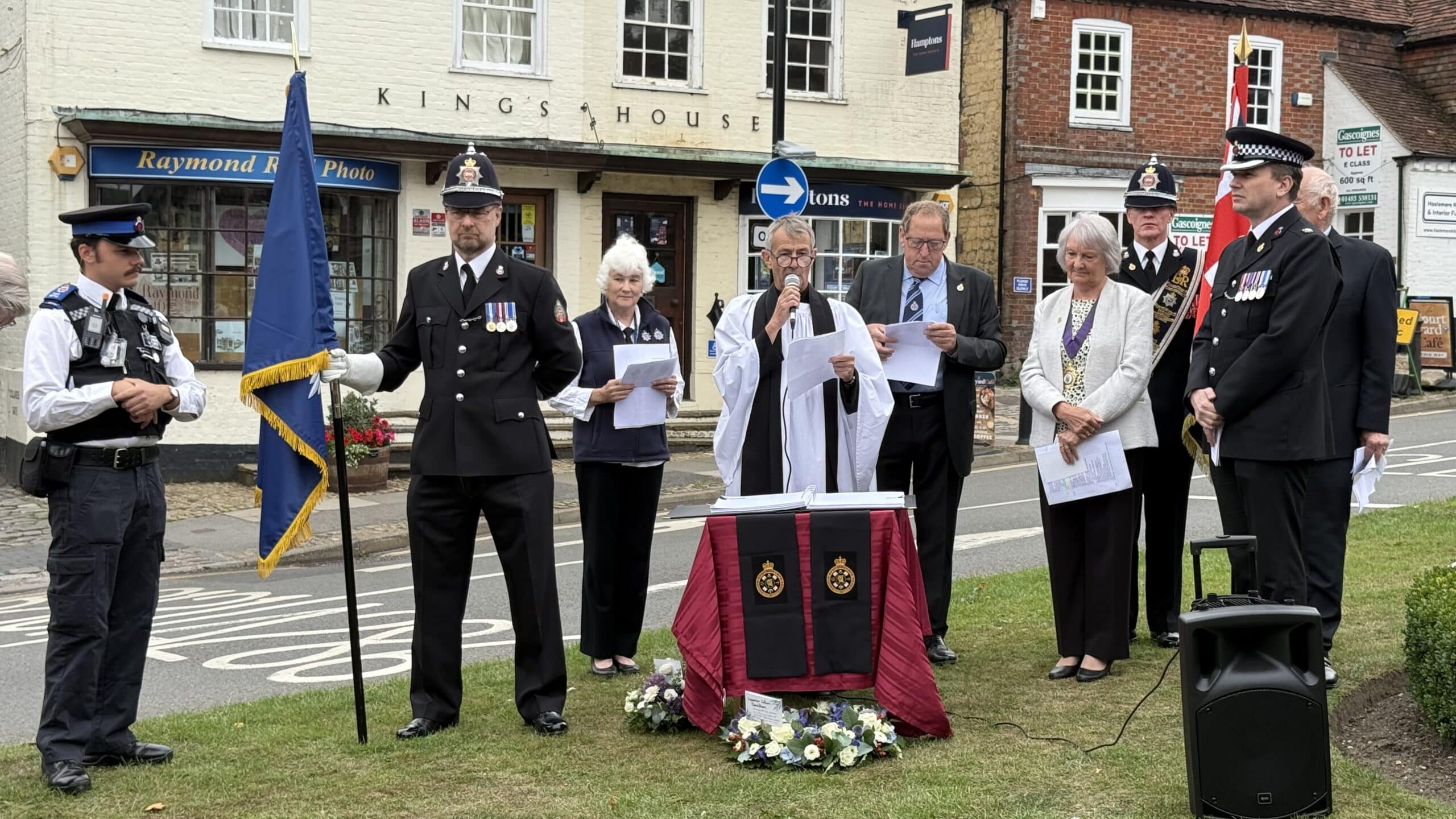 The picture shows a group of people standing on a green. They include people in formal police dress and a reverend, who is speaking into a microphone.