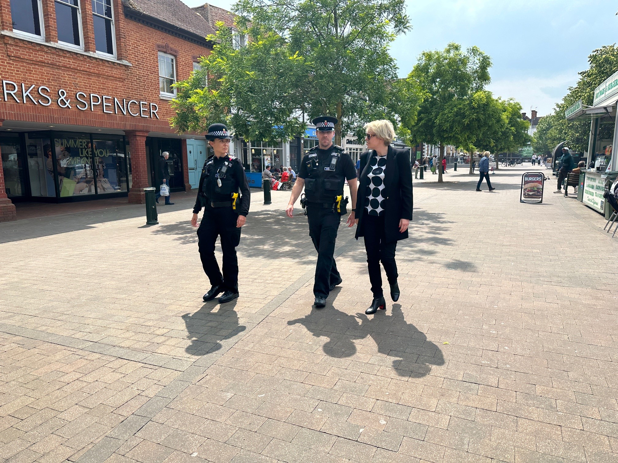 The image shows Police and Crime Commissioner Lisa Townsend walking through a town centre with two police officers in uniform. Lisa, who has blonde hair, stands on the right of the image. She wears a black jacket and trousers. The officers, one male and one female, are wearing their uniforms and police hats, and they are in the middle of a conversation with Lisa as they all walk past a branch of Marks and Spencer.