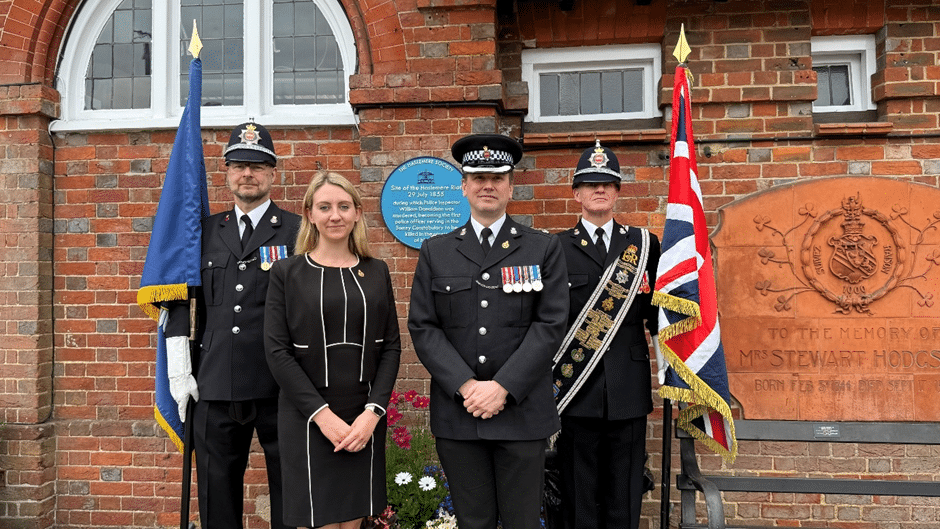 Image shows four people looking into the camera. On the left and right are two people in police uniform holding flags. In the middle is Deputy Commissioner Ellie Vesey-Thompson, a blonde woman in a black dress, and an officer with medals.