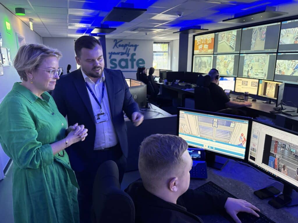 Two people stand and look at computer screens. One, a blonde woman in a green dress, is Commissioner Lisa Townsend. She stands next to a man in a suit, who wears a lanyard around his neck. A man is sitting at the computer with his back to the camera. In front of all three people pictured is a wall covered with screens, each of which shows CCTV images.