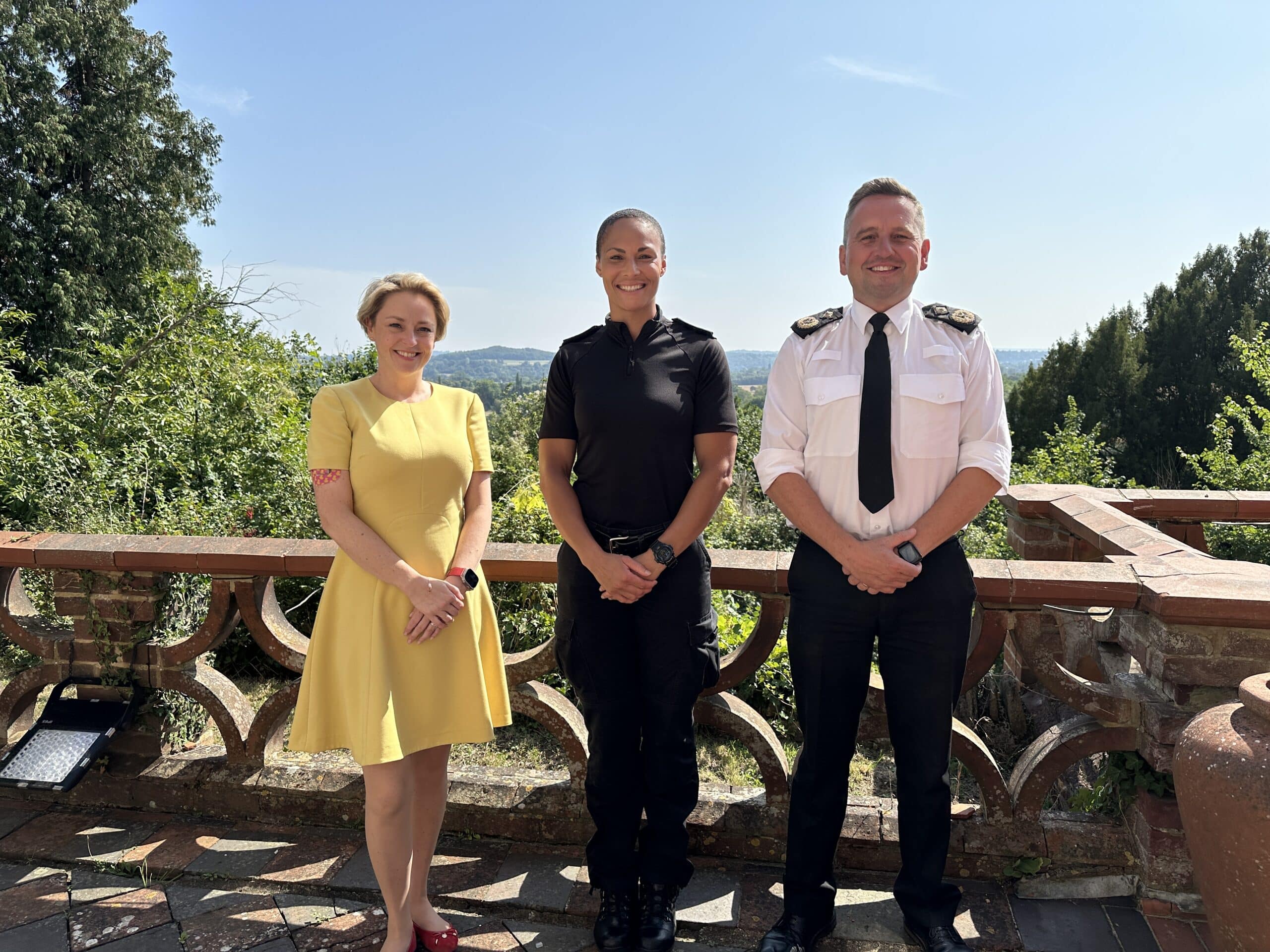Three people stand outside. All are facing the camera and smiling. On the left, wearing a yellow dress, is Police and Crime Commissioner Lisa Townsend, a blonde woman. To her left, in uniform, is Sergeant Candice Gill, and also in uniform, Surrey Police's Deputy Chief Constable Nev Kemp.