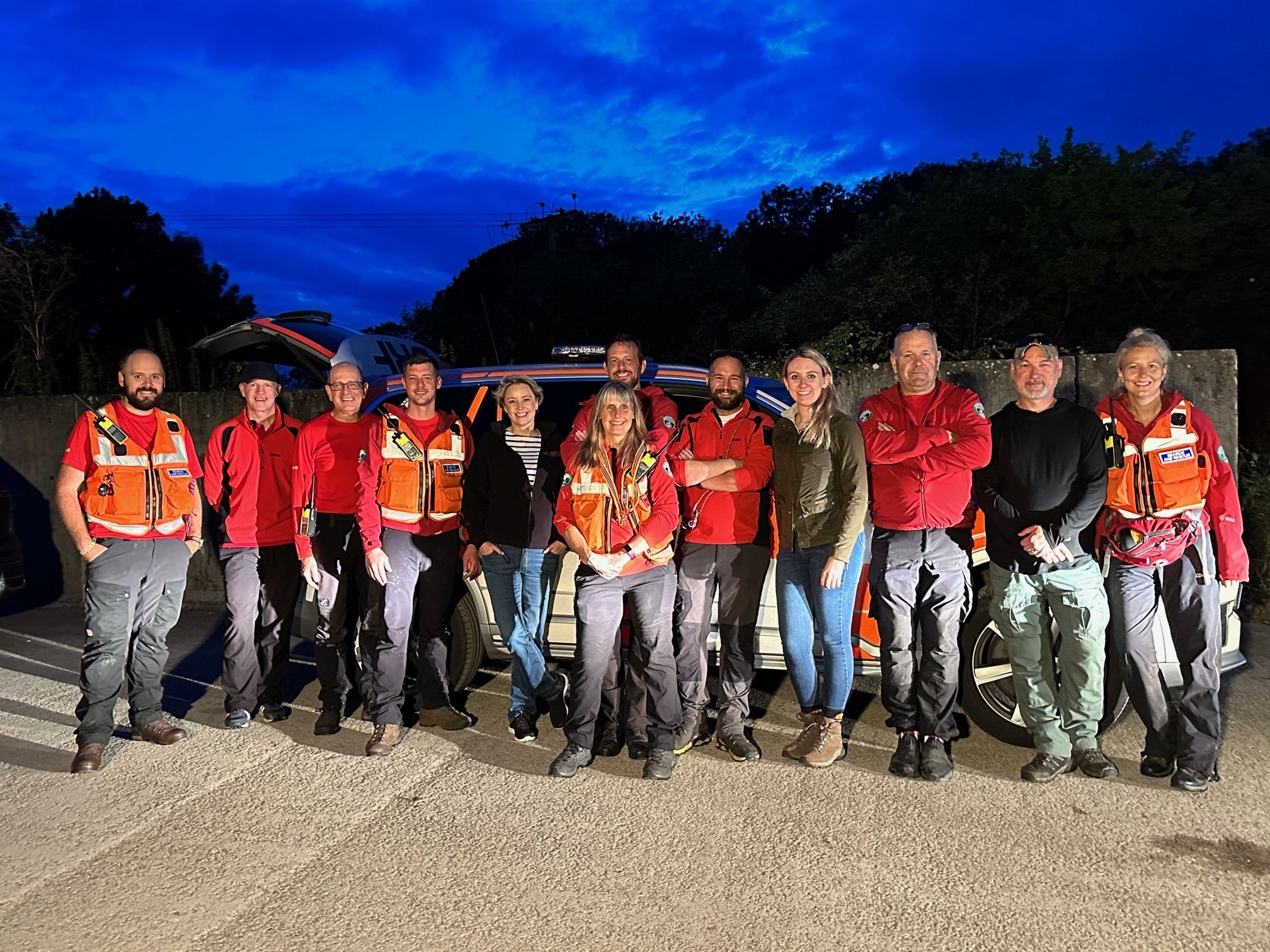 The image shows a group of people looking at the camera. Most are wearing a high-visibility uniform. They are the Surrey Search and Rescue volunteers. Also among the group are two blonde women wearing jeans and jackets. These women are Police and Crime Commissioner Lisa Townsend and Deputy Police and Crime Commissioner Ellie Vesey-Thompson. Everyone is looking at the camera and smiling. They are standing in a concrete car park, and it is night time.