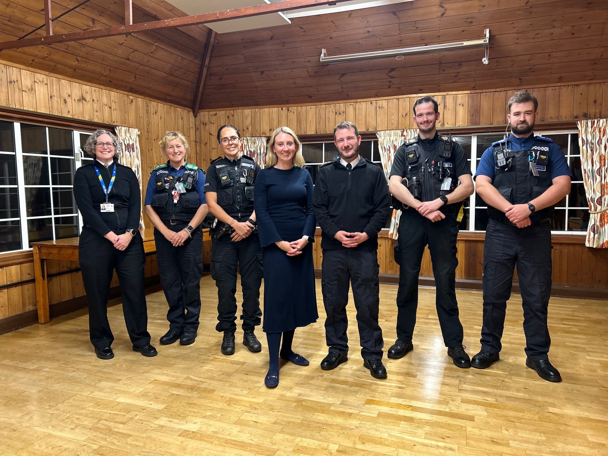 A group of people stand facing the camera. All have their hands clasped in front of them, and all are smiling. Five of the six people pictured are police officers and PCSOs in Surrey Police uniform. The last person pictured wears a blue dress.
