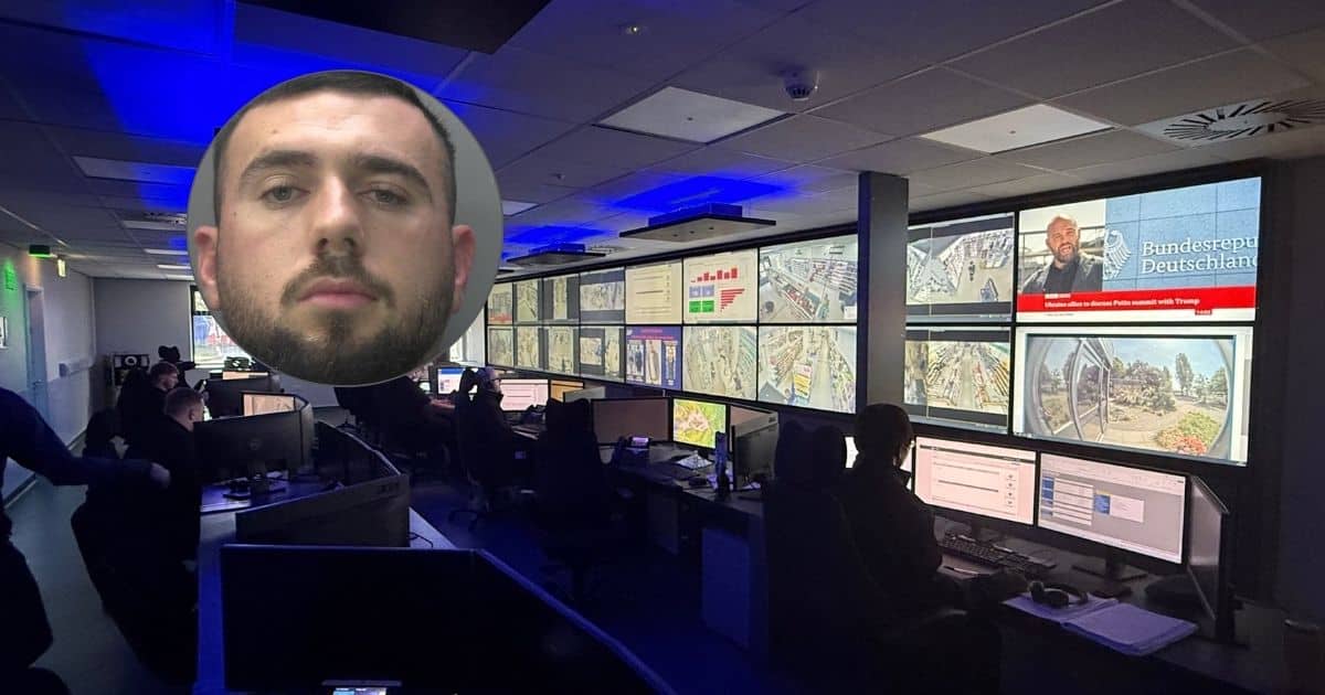 The image shows a dark room filled with computers and desks. Across the length of one wall is a bank of television screens, many of which show CCTV images. In the far left corner is a mugshot of a bearded man with dark hair, who looks directly into the camera.