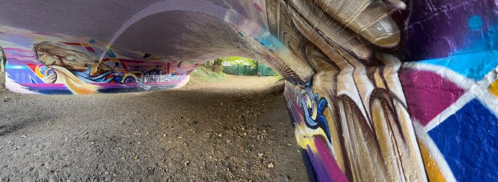 The image is a panoramic photo of a bridge over a public footpath. The bridge has been painted with images relating to Nescot College - it has bright colours and images from the college's history.