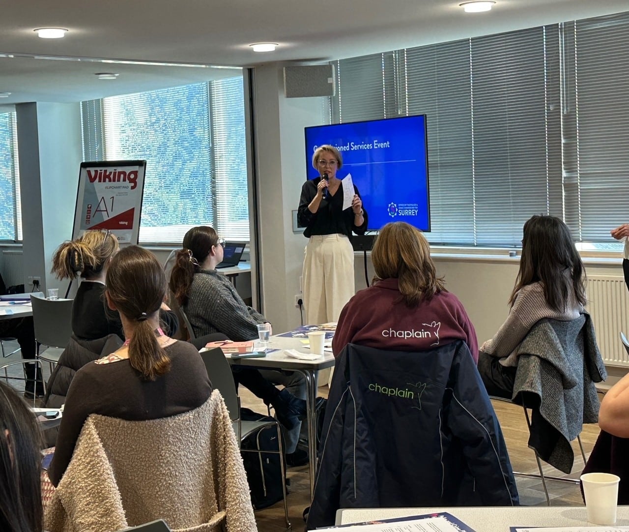 The image shows a group of people sitting at tables. They are facing away from the camera and looking at Police and Crime Commissioner Lisa Townsend, a blonde woman in white trousers and a darker-coloured top. Lisa is holding a microphone in one hand and a piece of paper in the other and is speaking to the audience.