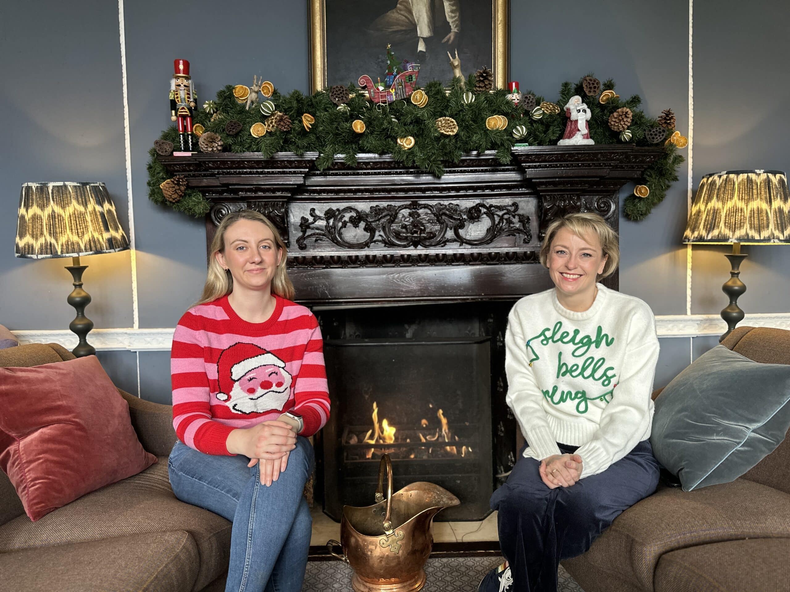 The image shows, on left, Deputy Police and Crime Commissioner Ellie Vesey-Thompson, a blonde woman wearing a pink and red Christmas jumper with Santa's face on, and on right, Police and Crime Commissioner Lisa Townsend, a blonde woman wearing a white Christmas jumper which reads 'sleigh bells ring'. Both women are sitting on sofas opposite each other with their hands clasped and legs folded. They are looking into the camera and smiling. Directly behind them is a blazing fireplace with Christmas decorations on the mantelpiece.