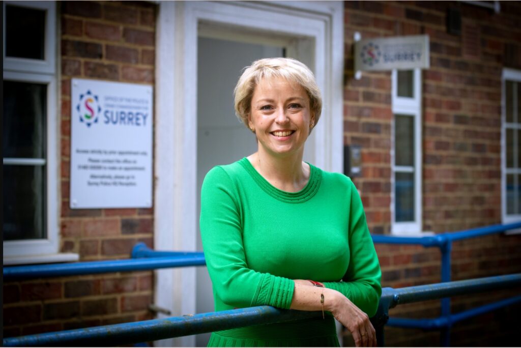 The image shows Police and Crime Commissioner Lisa Townsend, a blonde woman in a green dress, standing in front of a brick building. The building has a sign outside which reveals it to be the Office of the Police and Crime Commissioner. Lisa is leaning against a blue handrail, looking directly into the camera, and smiling.