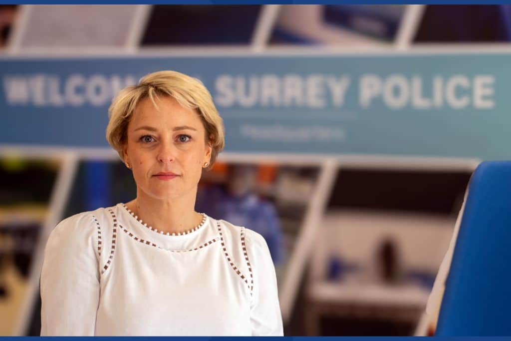 The image shows Police and Crime Commissioner Lisa Townsend, a blonde woman wearing a white shirt, standing in front of a large sign that reads 'Welcome to Surrey Police'. She is looking directly at the camera with a slight smile on her face.