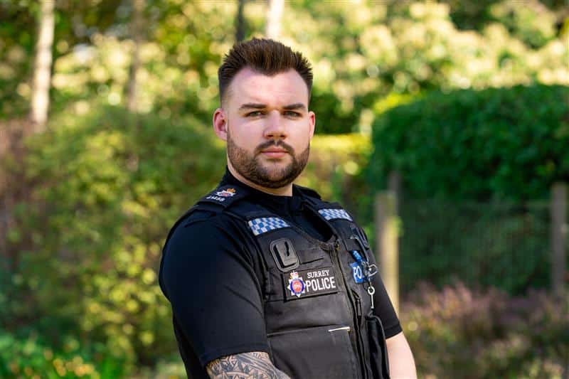 The image shows an officer in a stab vest looking straight at the camera with a stern expression. He is pictured against a leafy backdrop.