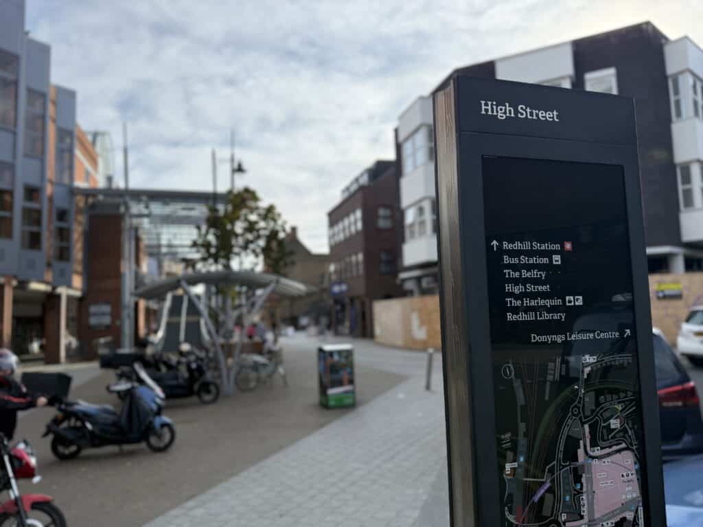The image shows Redhill High Street. It focuses on a sign that points out the train and bus stations, The Belfry shopping centre, the library and the theatre. Beyond the sign is an image of a street with a bike shelter and a moped visible.