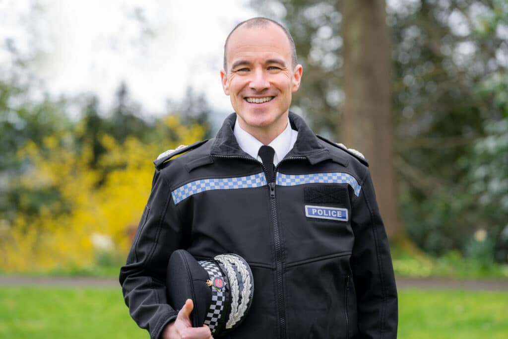The image shows Surrey Police's Chief Constable, Tim De Meyer. He wears full uniform, and carries his hand underneath one arm. He is looking directly into the camera and smiling.
