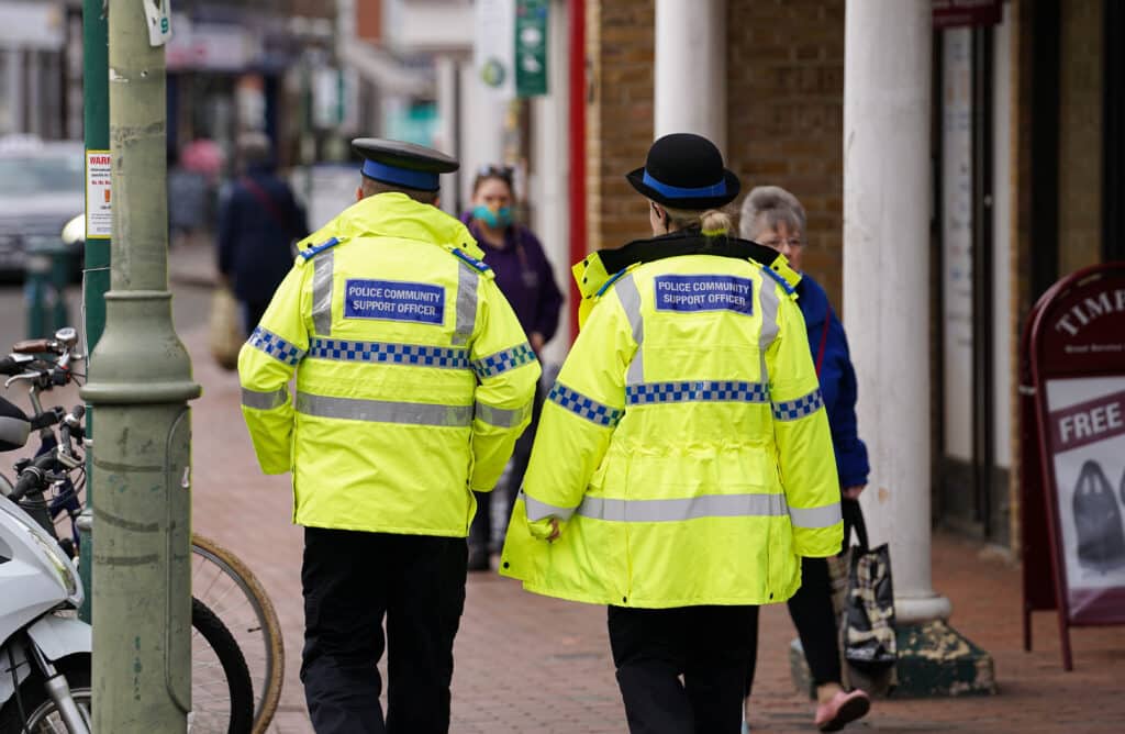 The image shows two Police Community Support Officers in high-visibility jackets and hats. They are walking down a busy high street as members of the public walk in the opposite direction.