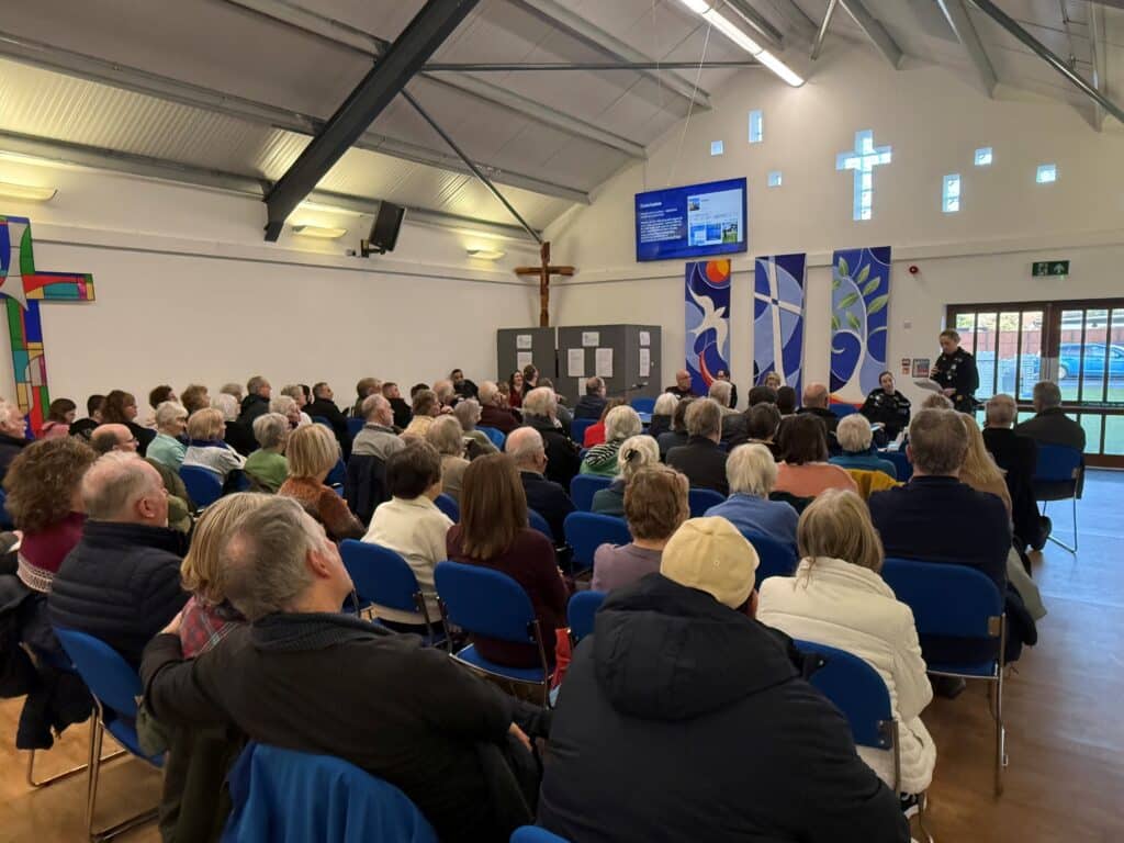 The image shows a large crowd of around 100 people sitting in a church hall. The audience is pictured from behind. They are facing a table, at which a police officer in uniform can be seen standing and speaking into a microphone. Above is a television screen showing a presentation.