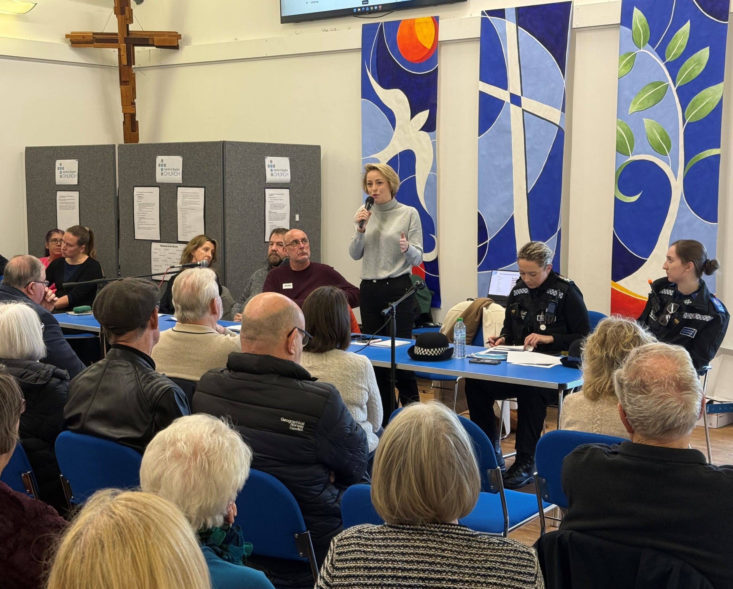 The image shows Police and Crime Commissioner Lisa Townsend, a blonde woman in a grey long-sleeved top, standing up at a table and speaking into a microphone. Seated at either side of her are police officers in uniform. In front of her is a large crowd who are sitting in chairs and pictured from behind.