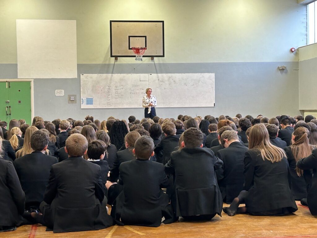 The image shows the Police and Crime Commissioner, a blonde woman in a shirt and trousers, standing at the front of a large group of schoolchildren. The children are seated on the floor of a gym with their backs to the camera. The Commissioner can be seen talking in front of a large whiteboard.
