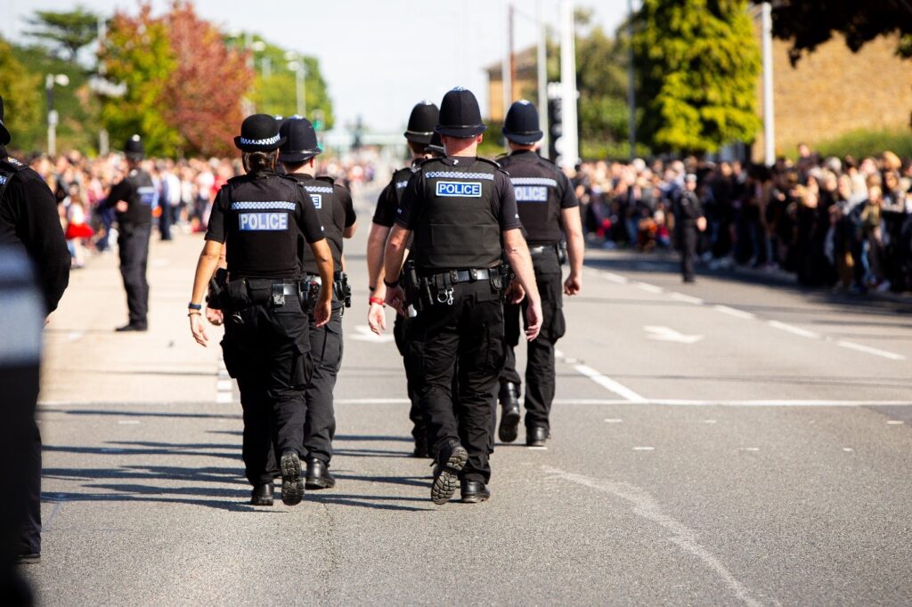 The image shows a large group of police officers in uniform walking along a main road between lines of crowds who have gathered at each side of the road. The faces of the crowd are blurred, with the officers' backs in sharp relief.