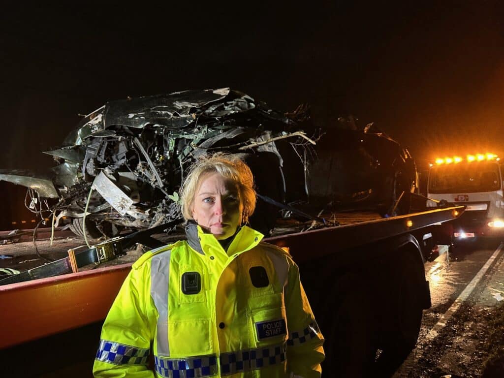 The photograph shows Police and Crime Commissioner Lisa Townsend, a blonde woman wearing a high-visibility jacket. She is standing in front of the wreckage of a car which has been lifted onto a low-loader. She has a stern expression on her face and is looking directly into the camera.