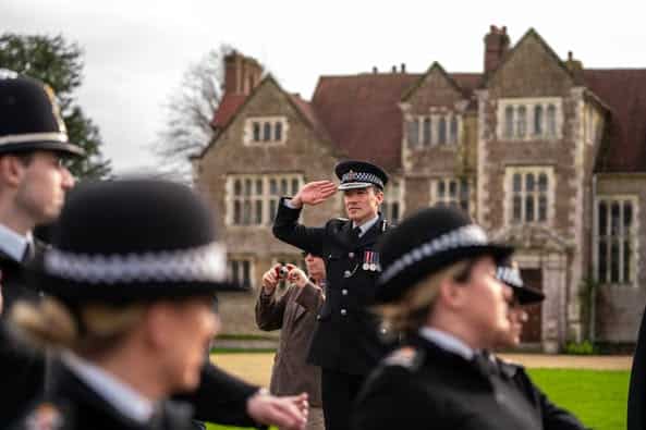 The image shows Surrey Police's Chief Constable, Tim De Meyer, saluting in front of an old stone property. In front of the Chief Constable are marching officers wearing full uniform. The image was taken at a 'passing out parade', which celebrates new recruits.
