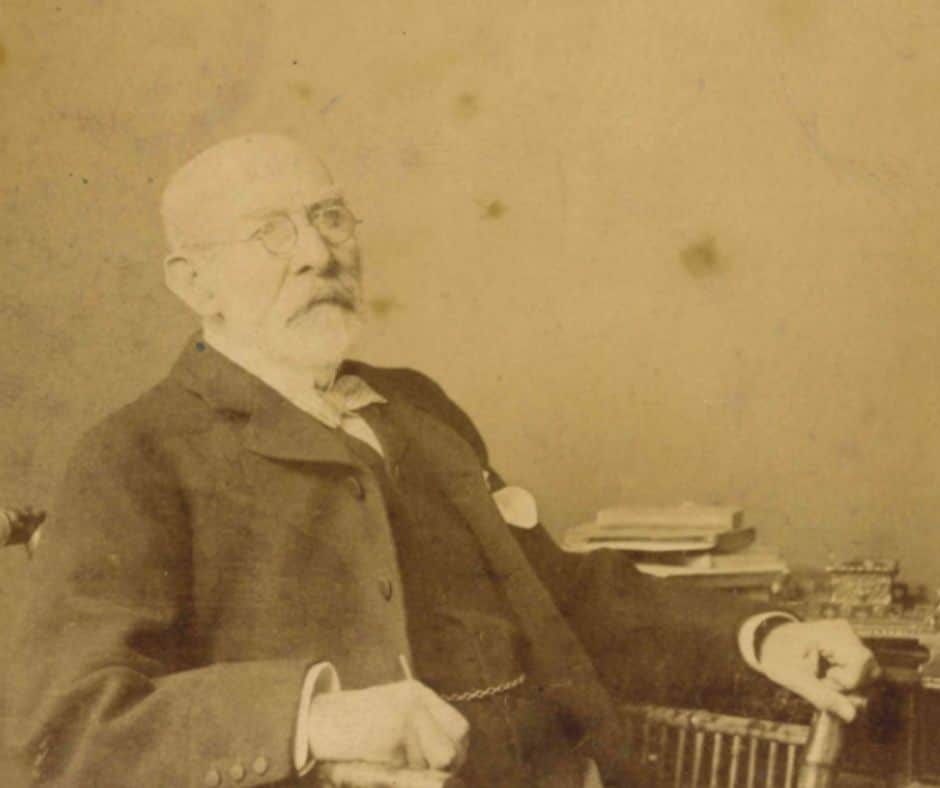 The image is a sepia-toned photograph. It shows a man with white hair, wire-framed glasses and a beard sitting in a chair at a desk. His arms rest on the arms of the chair, and he wears a black jacket and waistcoat. He has a serious expression on his face and is looking slightly to his left.