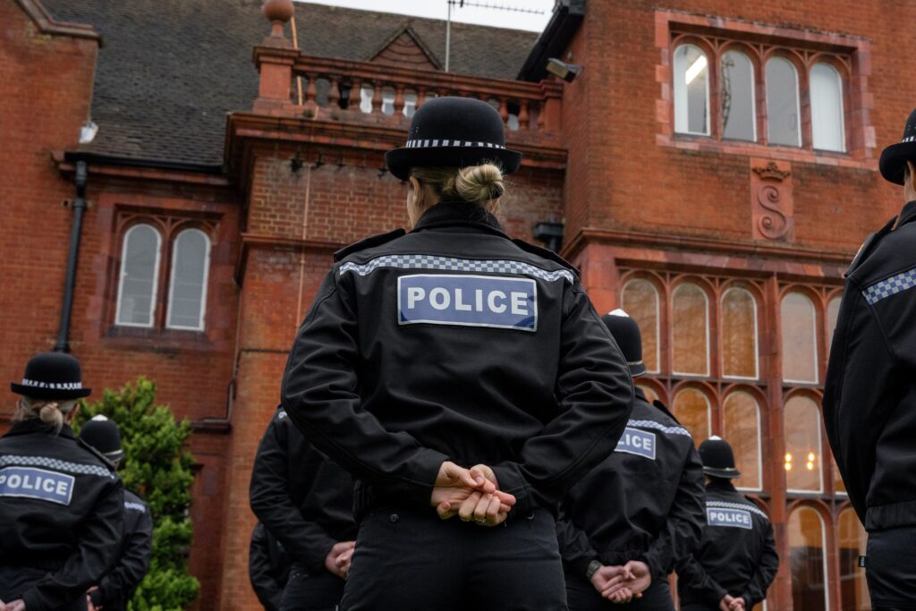 The image shows police officers in uniform standing in rows. They are photographed from behind, with the 'police' emblem on their jackets visible. Their hands are clasped behind their backs and they are facing a large, red-brick building, which can be found at Surrey Police's Mount Browne HQ in Guildford.