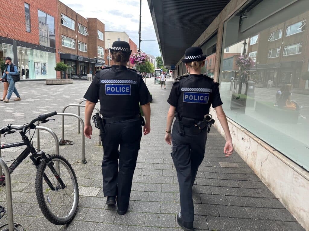 The image shows two police officers in uniform walking through a street in Camberley. The officers are pictured from behind as they stroll through the street.