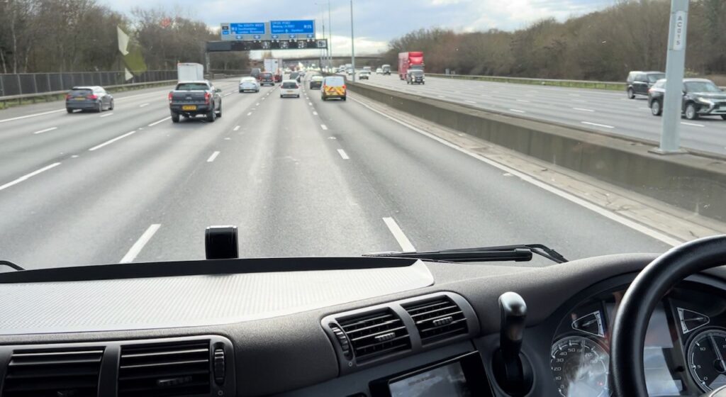 The image shows a stretch of the M25 taken from the cab of an HGV lorry. Ahead is a gantry with blue signs above it. There are cars in the carriageway.
