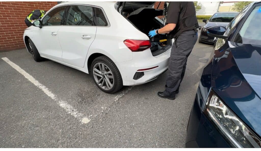 The image shows an officer in black and wearing latex gloves looking through the boot of a white car. The officer's face is not visible, but their hands and body can be seen as they look through the vehicle.