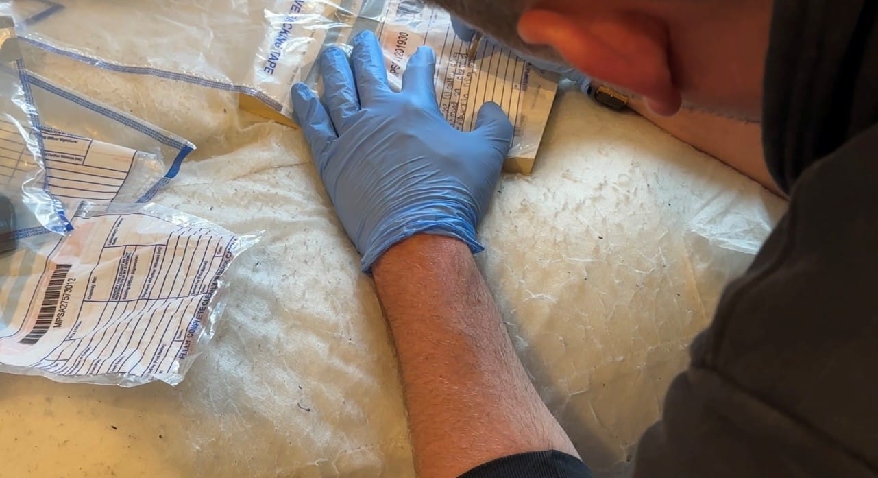 The image shows a Crime Scene Investigator with blue latex gloves on. He is bent over and writing on an evidence bag with a pen. 