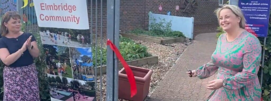 The image shows Police and Crime Commissioner Lisa Townsend, who wears a purple and green dress, smiling broadly with a pair of scissors in her hand as a ribbon falls away. To her right is a sign that reads 'Elmbridge community', while another woman is seen applauding. The image was taken at the opening of the St John Community Garden.