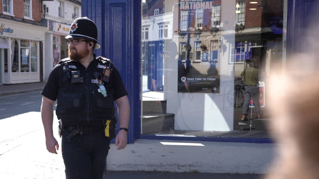 Strengthening relationships The image shows a police officer in full uniform walking along a street. He is looking away from the camera and smiling slightly. Behind him is a branch of bookmakers Coral.