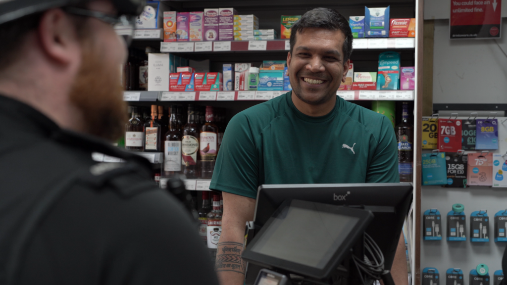 The image shows a man smiling at a police officer from behind a till. Only the back of the police officer is visible, although his uniform and hat can be seen. The second man in the image is smiling broadly at the officer. Behind him are shelves which have bottles of alcohol and other items on them.