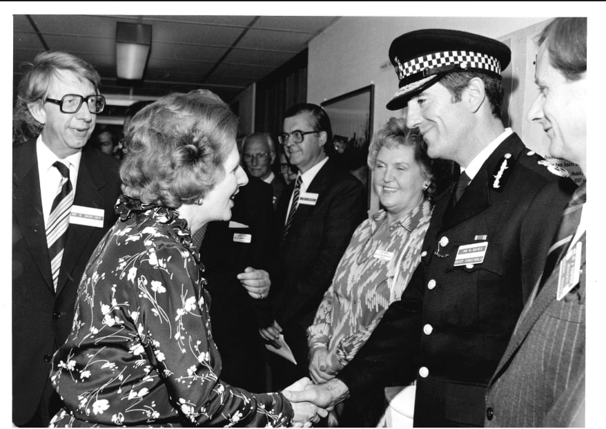 The black and white image shows a large crowd of people standing in a hallway. The image focuses on two people - an officer in full uniform, and a woman with brown hair in a patterned top. They are shaking hands and smiling at each other. The woman is then-Prime Minister Margaret Thatcher, and the man in uniform is former Surrey Police Chief Constable Brian Hayes.