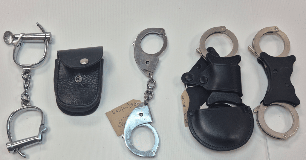 The image shows sets of handcuffs lined up on a table. They range from historic to the modern police handcuffs we recognise today.