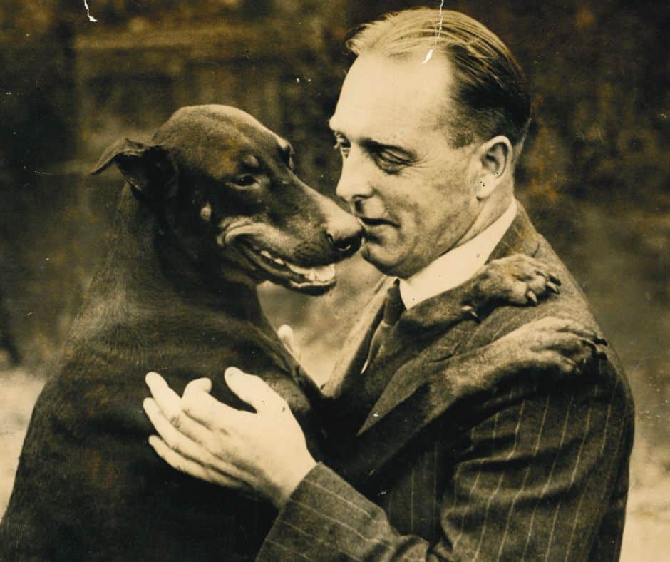 The image, which is in sepia tones, shows a large dog on the left and a man on the right. The man wears a suit, and the dog's paw lies on his shoulder. The man has his hand on the dog, and their faces are close together in an affectionate pose.