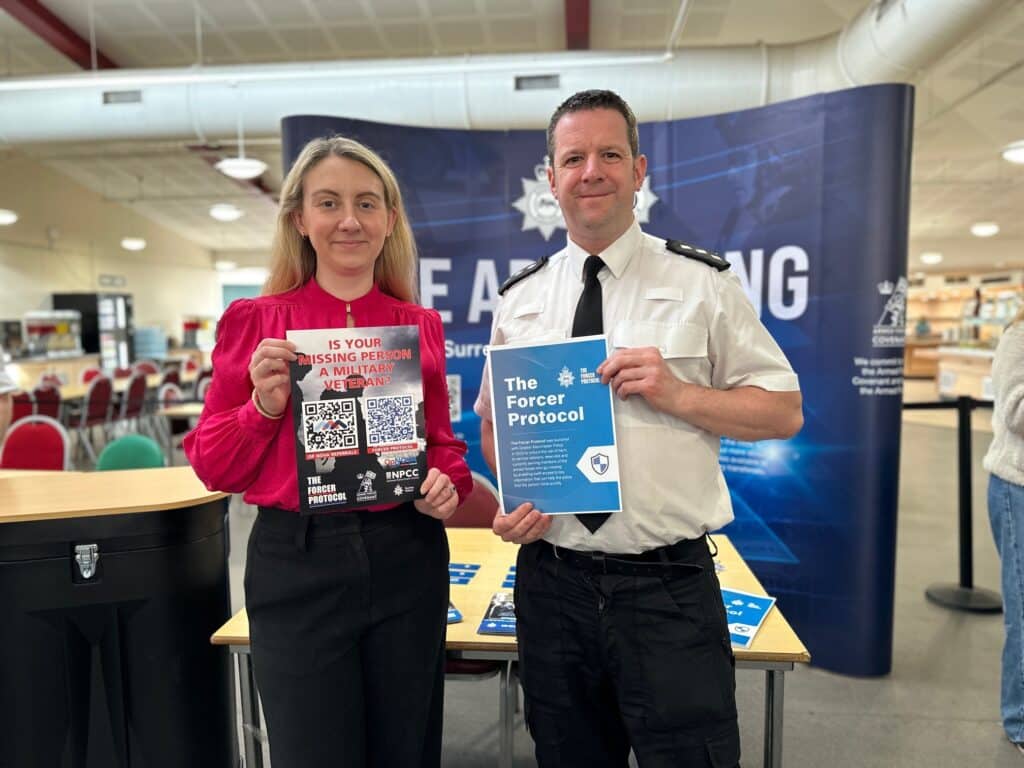 Two people hold posters and smile directly at the camera. On the left is Deputy Police and Crime Commissioner Ellie Vesey-Thompson, a blonde woman in a pink blouse and black trousers. On the right is Chief Inspector Tom Renwick, who wears a shirt and tie. In their hands are posters advertising the Forcer Protocol, which aims to help police find missing veterans promptly.
