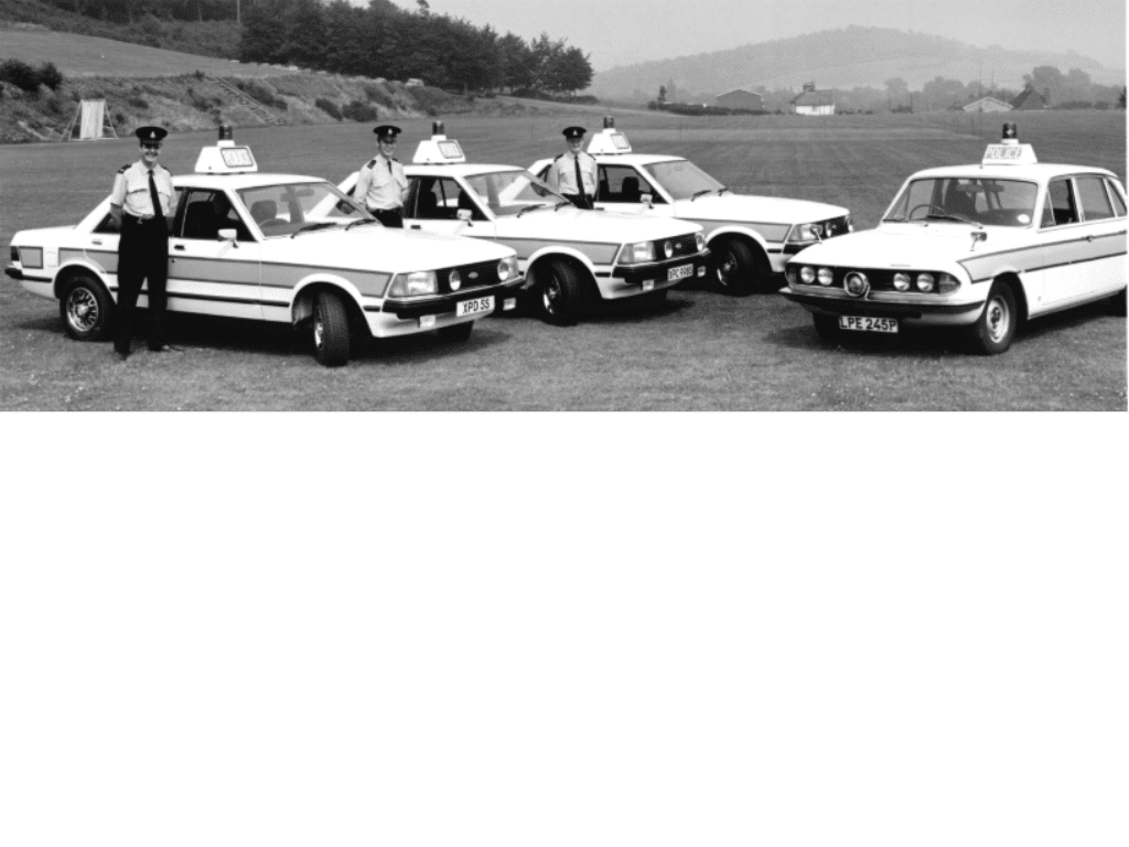 Black and white image of 4 white Ford Granadas parked alongside each other in a field.  It is a scenic location with hills in background.  On the roof of each car is a police logo and blue siren light on top.  There are three police officers in uniform standing beside the cars.