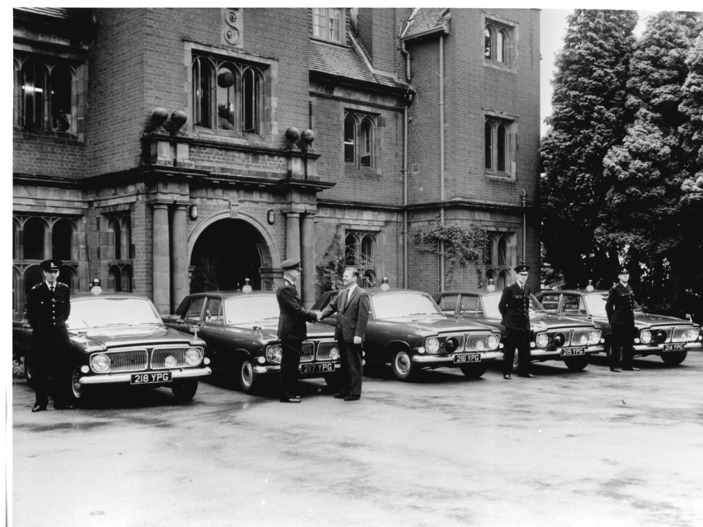 Black and white image of five Ford Zephyrs parked outside the Old House at Mount Browne.  Each vehicle is dark in colour and has a blue siren light on its roof.  There are three officers in uniform standing in front of the cars.  One gentleman is shaking hands with another police officer.